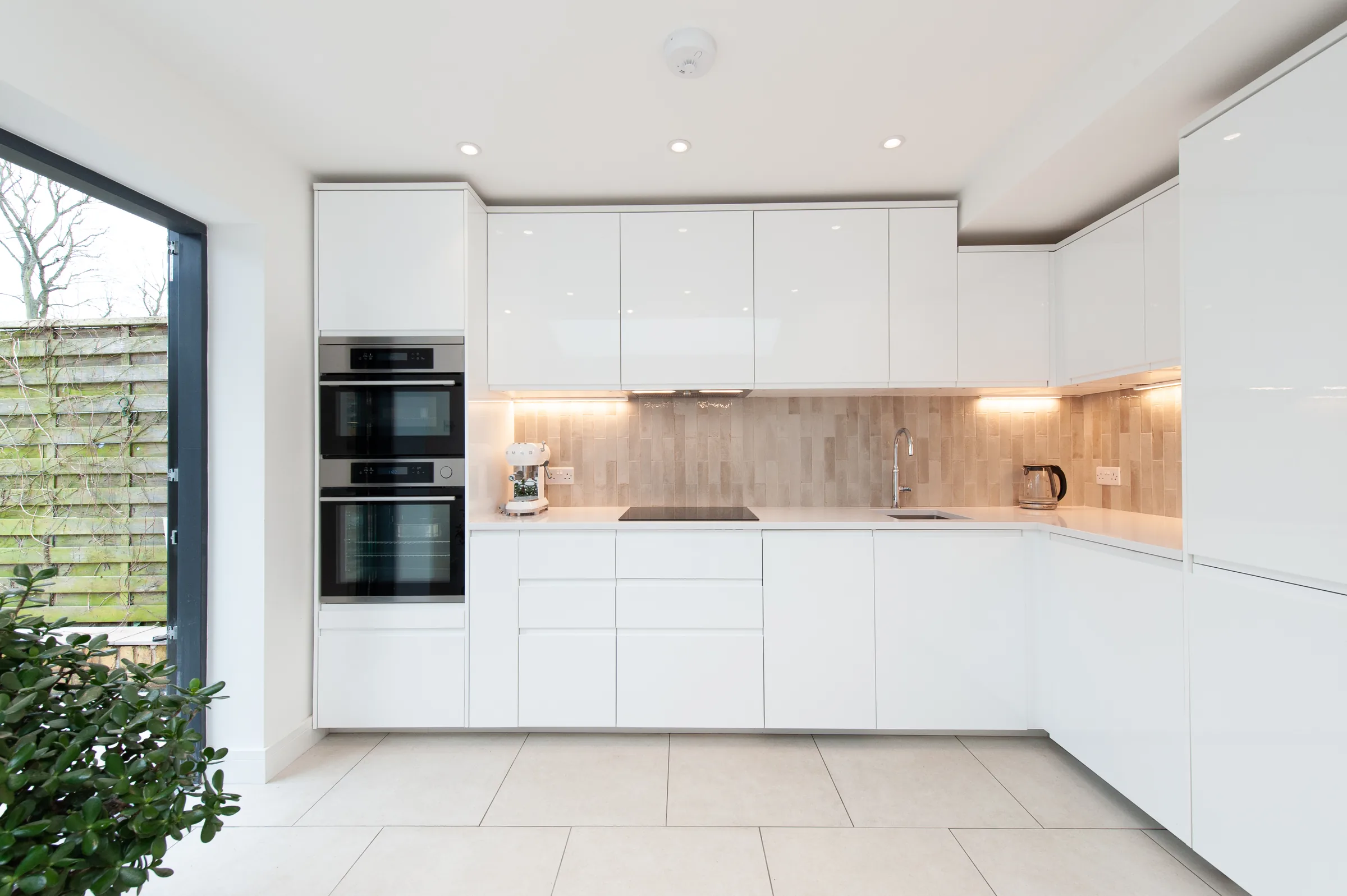 Bright white gloss kitchen with a textured tile splashback and full-height glazing onto the garden